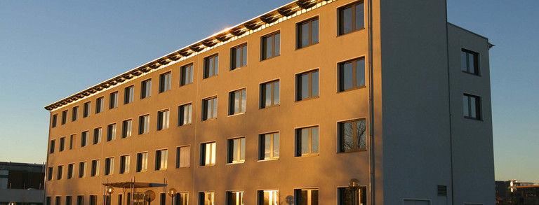 Gebäude E-Technik: Ein hohes weißes Gebäude, das von der Sonne angestrahlt wird. Vor dem Gebäude eine Rampe und über dem Eingang ein Vordach.