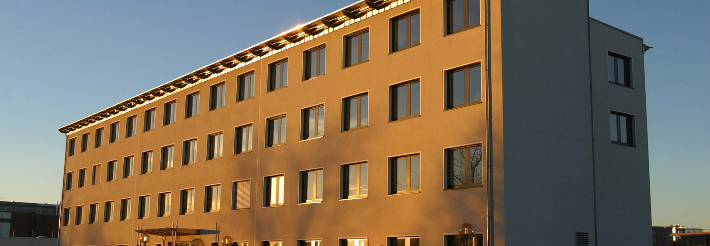 Gebäude E-Technik: Ein hohes weißes Gebäude, das von der Sonne angestrahlt wird. Vor dem Gebäude eine Rampe und über dem Eingang ein Vordach.